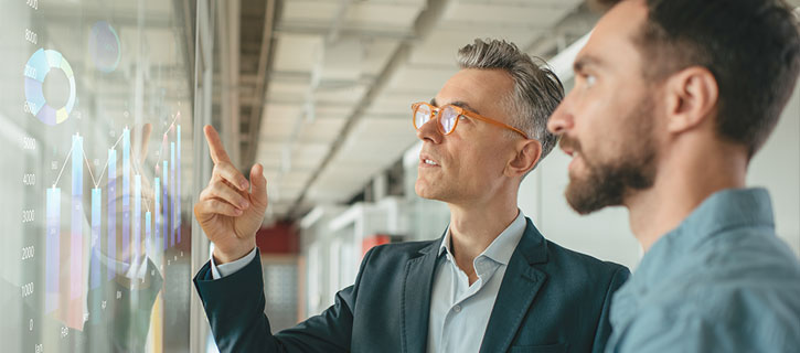 Zwei Geschäftsmänner stehen vor einem großen Bildschirm, der wichtige Kennzahlen anzeigt. Die Daten wurden im Rahmen von Industrie 4.0 über das Internet der Dinge generiert.