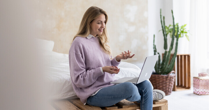 Eine junge Frau sitzt entspannt auf einem Bett und führt lächelnd ein Gespräch über ihren Laptop.
